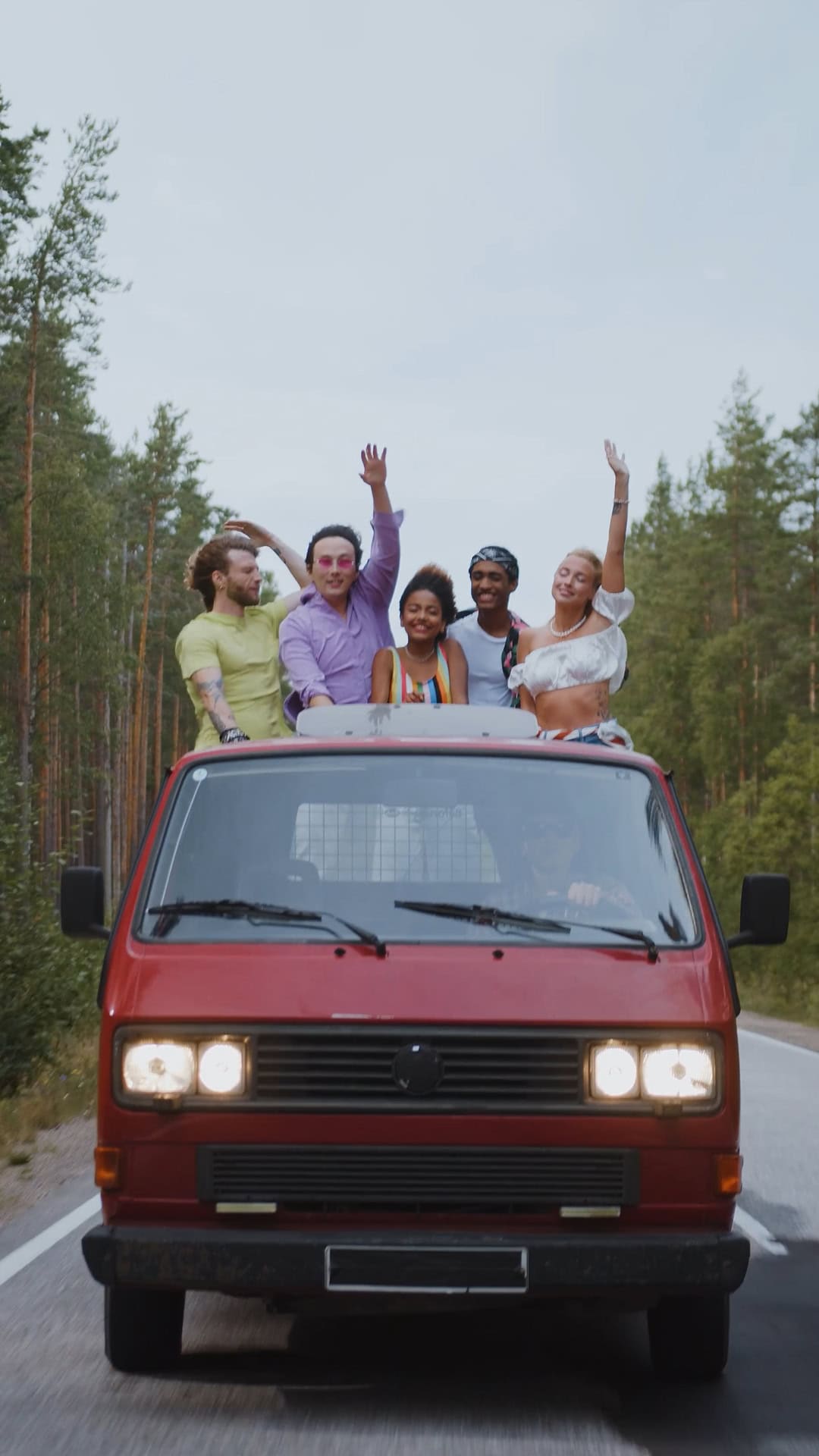 Eine Gruppe von Freunden jubelt vom Dach eines VW-Busses aus, der eine Straße hinunterfährt, und hält einen Moment der Freude und des Abenteuers fest