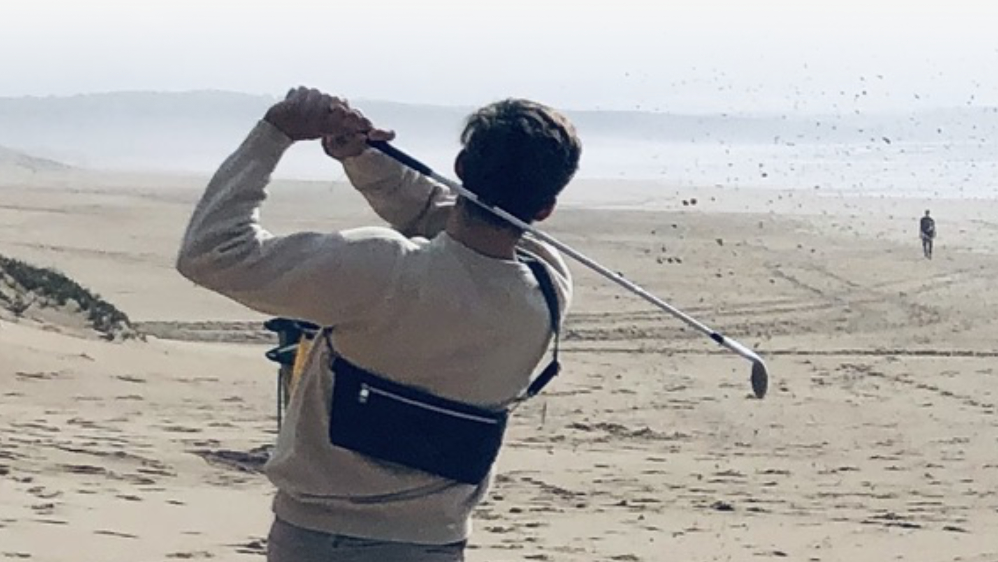 Golfer wearing Manufabo Belt Bag, swinging at a ball on the beach, symbolizing the outlet sale of bags flying off the shelf and into the sunset