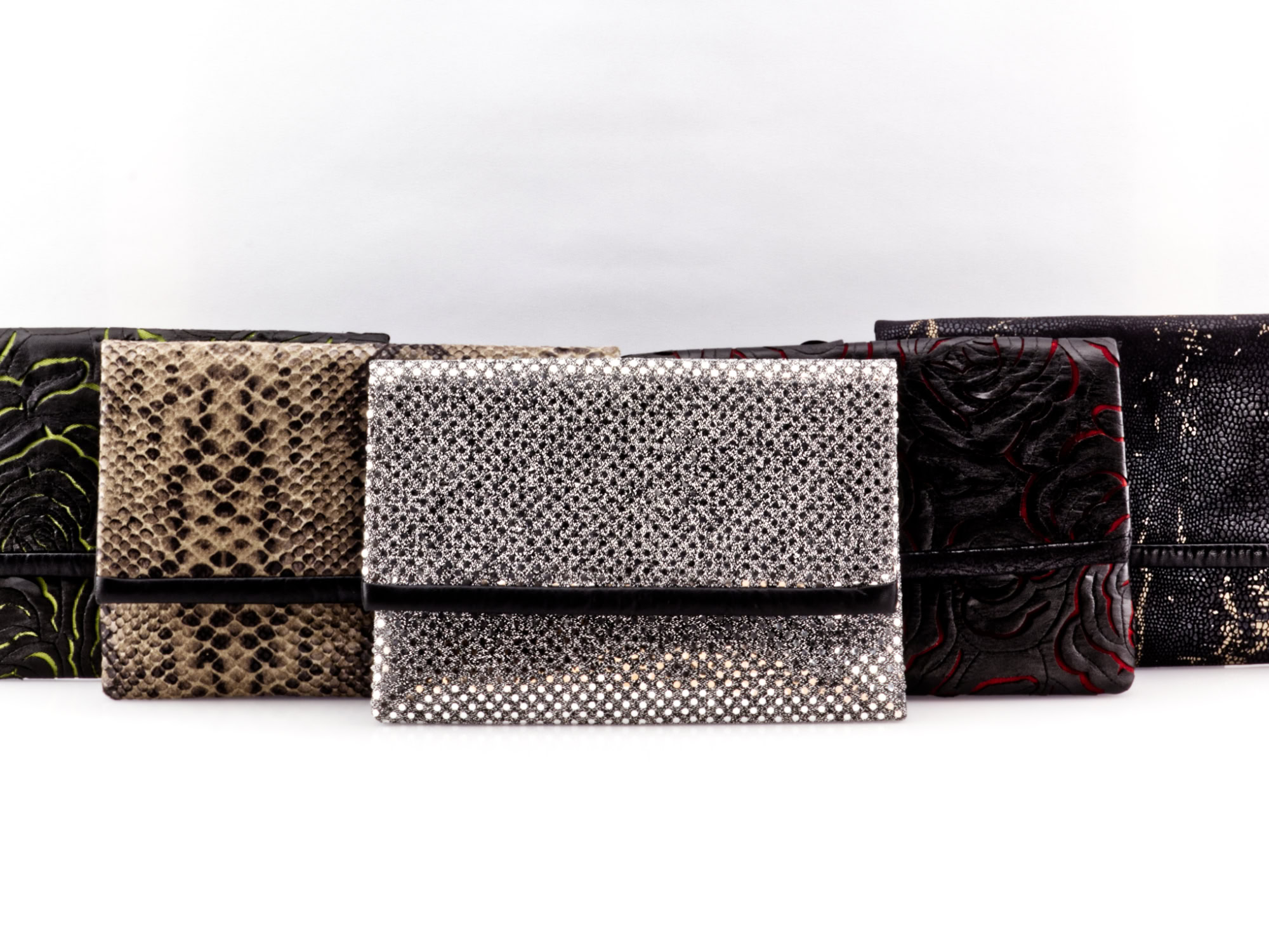 Five Manufabo clutches arranged in a V-shape: silver sequins, black reptile, rose patterns in green and red, and snake leather