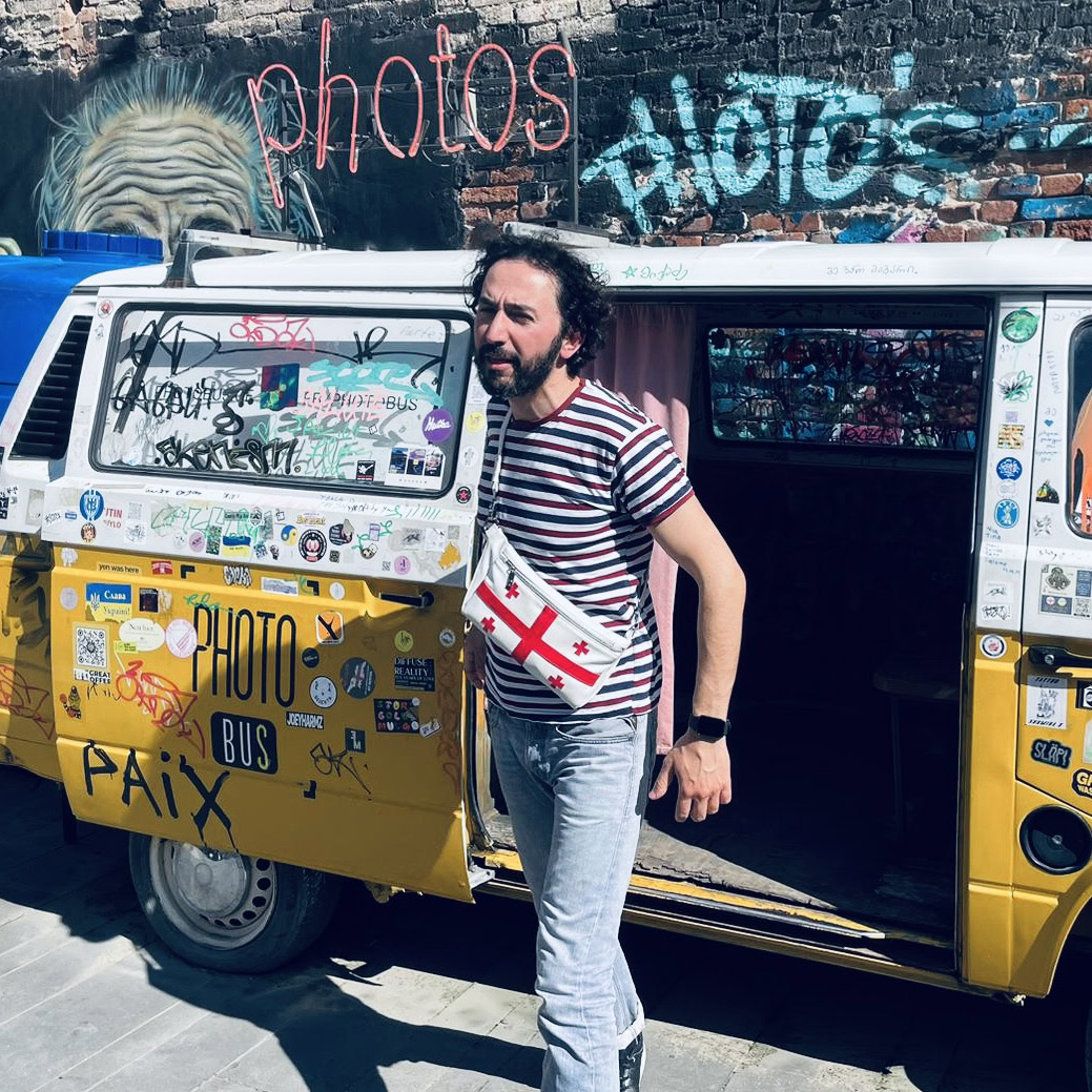 Proud Georgian Stud with St George Cross Bag in Red and white stepping out of vw camper photo bus in Tbilisi with street art