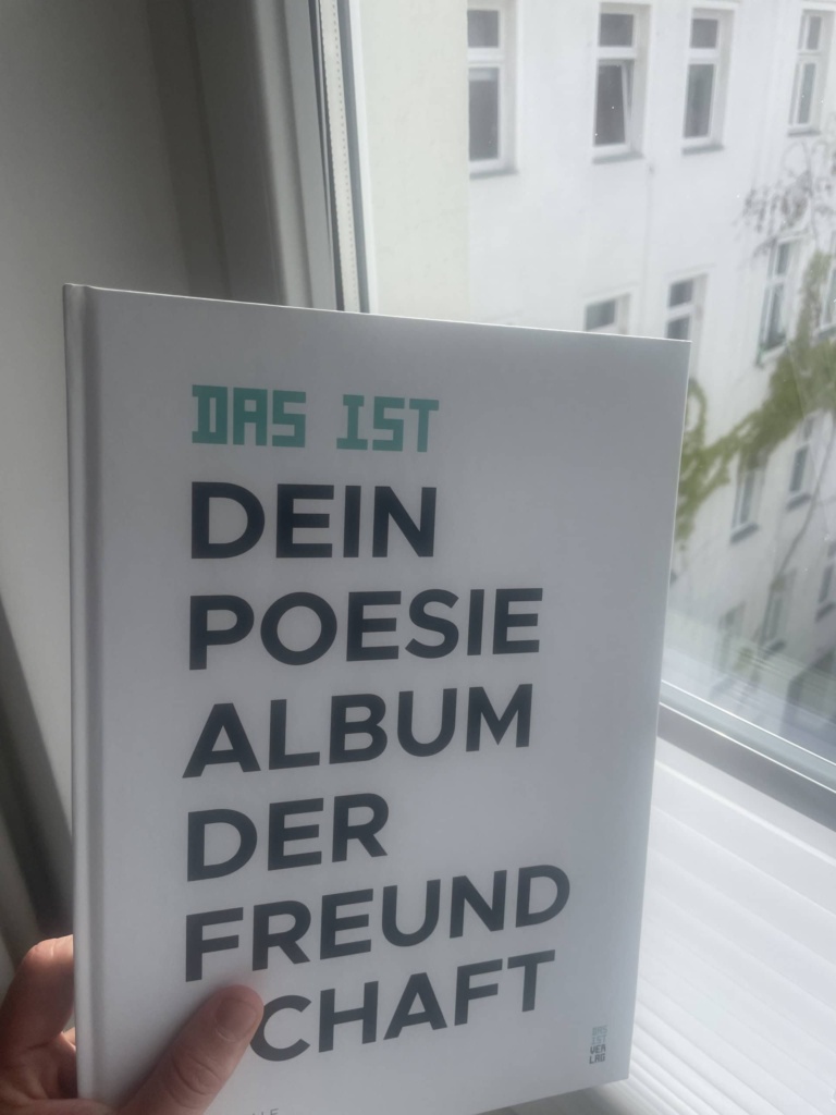 Das weiße Cover des Freundebuchs, vor ein Fenster gehalten, dahinter die Fassade eines Altstadt-Innenhofs